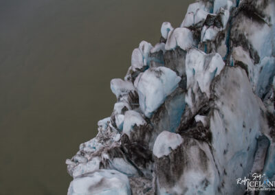 A close-up view of a glacier, showcasing various ice formations with smooth, rounded edges and patches of dark rock interspersed among the bright blue and white ice. The background features a calm, murky body of water, reflecting the glacier's colors. The texture of the ice appears rugged in some areas, while others have a polished look due to melting.