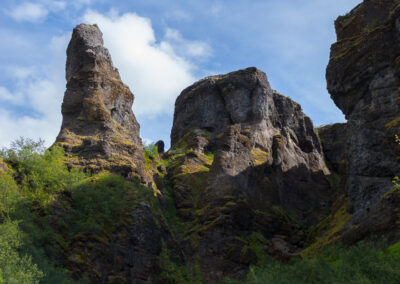 A dramatic landscape featuring towering rock formations against a blue sky, partially covered in clouds. The rocks exhibit rugged textures and are surrounded by lush greenery at their base, with small shrubs and plants peeking through the rocky terrain. The scene captures the natural beauty and geological characteristics of the area.