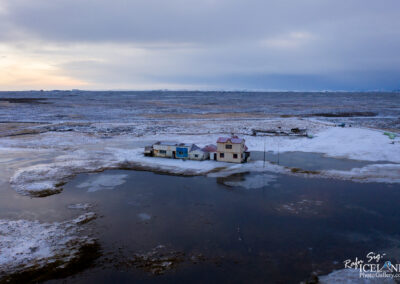 A remote winter landscape featuring two houses, one blue and one white with red accents, situated on a small patch of land surrounded by icy water. The terrain is predominantly covered in snow, with patches of dark earth visible. In the background, a vast flat area extends towards the horizon, under a cloudy sky that hints at dusk, casting a soft, muted light over the scene. Sparse vegetation and a few distant structures are visible, emphasizing the isolation of the location.
