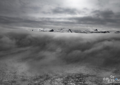 A dramatic black and white landscape featuring a layer of dense clouds obscuring the lower section of a glacier or ice field. Above the clouds, snow-capped mountain peaks emerge, silhouetted against a soft gray sky with scattered clouds, creating a moody and ethereal atmosphere. The intricate patterns of the glacier are visible near the bottom of the image, adding depth to the scene.