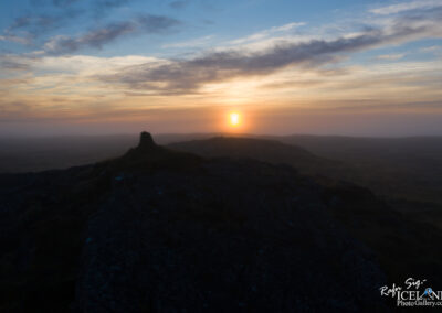 A panoramic view of a misty landscape at sunset, featuring a low mound on the left side of the image. The sun is setting behind distant hills, casting a warm glow that contrasts with the cool tones of the surrounding sky and mist. Wispy clouds scatter across the sky, adding depth to the horizon. The rocky terrain is mostly dark, hinting at the natural features of the land.