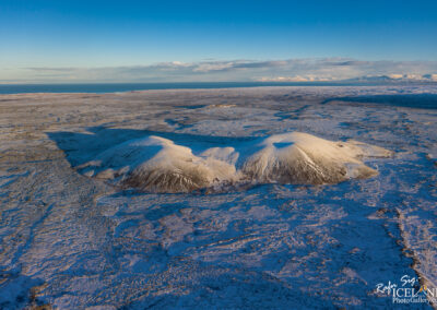 An aerial view of two snow-capped volcanic mountains set against a vast, icy landscape. The terrain is mostly flat with a mix of blue and brown hues, and the ocean is visible in the background under a clear blue sky with scattered clouds. The scene conveys a sense of serenity and stark natural beauty.