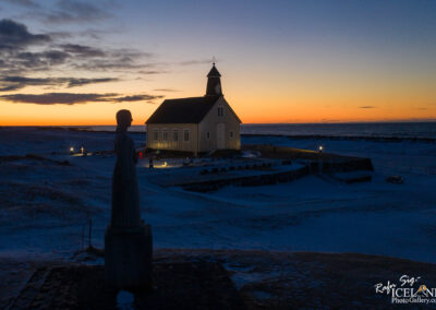 A peaceful winter landscape at sunset featuring a small, white church with a black roof near the ocean. In the foreground, a statue of a woman stands in profile, casting a dark silhouette against the colorful sky. The ground is covered in snow, and several illuminated crosses are visible in the surrounding area, creating a serene atmosphere as the day transitions to night.