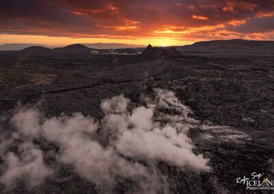 A dramatic aerial view of a volcanic landscape at sunset, featuring dark, textured lava fields interspersed with wisps of steam and smoke rising from the ground. In the background, distant hills are silhouetted against a vibrant sky painted with shades of orange, red, and purple as the sun sets, casting a warm glow over the rugged terrain.