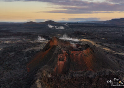 Aerial view of a volcanic landscape with a prominent volcanic cone in the foreground. The cone has reddish and dark rocky textures, with visible steam rising from fissures. The landscape is predominantly black lava rock, spreading out towards the horizon where gentle hills and mountains can be seen under a soft, muted sky during twilight. The overall atmosphere is dramatic and rugged, characteristic of volcanic terrain.