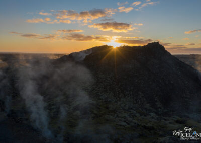 A stunning sunset over a volcanic landscape, with the sun's rays breaking through clouds and illuminating a rocky peak. Wisps of steam rise from the terrain, enhancing the dramatic and mysterious atmosphere of the scene. The horizon stretches into the distance, blending the earth and sky in soft, warm hues.