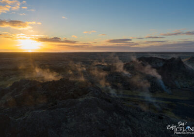 A stunning aerial view of a volcanic landscape at sunset. The scene displays rugged black lava rock formations, with steam rising in various places, set against a sky filled with soft clouds illuminated by warm orange and yellow hues as the sun sets on the horizon. The vast, barren terrain stretches into the distance, showcasing the unique geological features of the area.