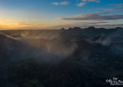 A panoramic view of a rugged volcanic landscape at sunset, featuring dark, rocky terrain with low-lying mist and steam rising from the surface. In the background, silhouettes of distant peaks are visible against a colorful sky filled with soft clouds, transitioning from orange to blue as the sun sets on the horizon.