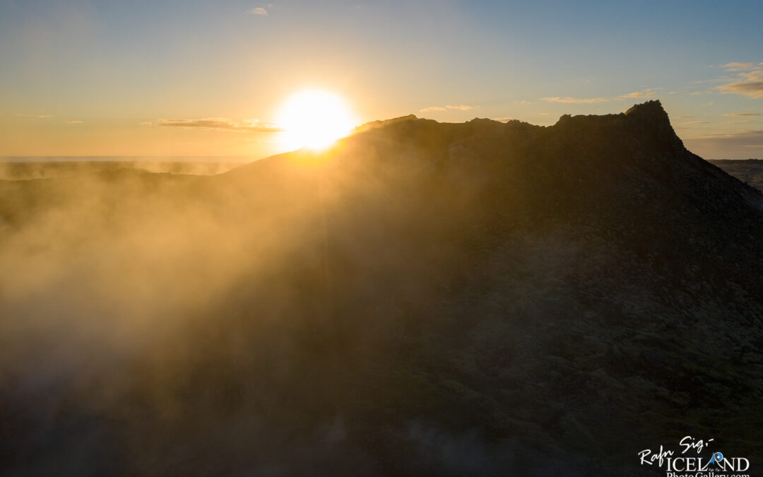 A dramatic landscape at sunrise, featuring a mountain silhouette against a bright sun emerging from behind the peak. Soft mist envelops the lower part of the mountain, creating a serene and ethereal atmosphere. The sky is partly cloudy, with soft hues of orange and blue blending together in the background.