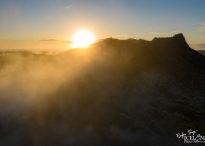 A dramatic landscape at sunrise, featuring a mountain silhouette against a bright sun emerging from behind the peak. Soft mist envelops the lower part of the mountain, creating a serene and ethereal atmosphere. The sky is partly cloudy, with soft hues of orange and blue blending together in the background.