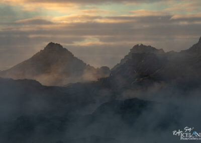 A dramatic landscape featuring rugged, dark rock formations partially shrouded in mist, set against a soft, cloudy sky with subtle hues of blue and gold as the sun rises or sets. The uneven peaks create a sense of depth and mystery, enhanced by the ethereal fog that swirls around the base of the rocks.