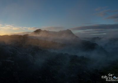 A misty landscape at sunrise featuring rugged hills and rocky terrain. The low light creates a hazy atmosphere, with soft clouds in the sky and steam rising from the ground, suggesting geothermal activity. The overall mood is tranquil yet otherworldly, capturing the natural beauty of a volcanic region.