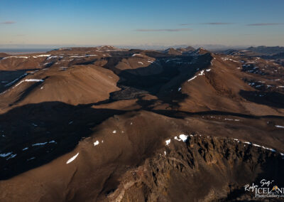 Aerial view of a mountainous landscape featuring a series of rugged hills and valleys. The terrain is predominantly brown and gray with patches of white snow visible on the peaks and ridges. In the background, additional mountain ranges can be seen against a clear blue sky. The play of light and shadow accentuates the contours of the land, creating a dramatic and picturesque natural scene.
