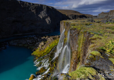 A stunning waterfall cascading down a moss-covered cliff into a turquoise river below, surrounded by rugged rocky terrain and steep cliffs in the background. The scene is set under a partly cloudy sky, providing a dramatic contrast between light and shadow across the landscape.