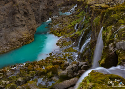 A scenic view of a river flowing between rocky cliffs, featuring vibrant turquoise water. The landscape is adorned with patches of green moss and small waterfalls cascading down the rocks into the river below. The overall atmosphere is serene and natural, highlighting the beauty of the surrounding environment.