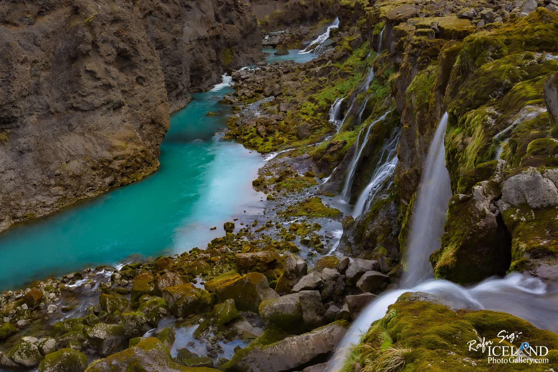 A scenic view of a river flowing between rocky cliffs, featuring vibrant turquoise water. The landscape is adorned with patches of green moss and small waterfalls cascading down the rocks into the river below. The overall atmosphere is serene and natural, highlighting the beauty of the surrounding environment.