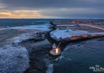 An aerial view of a coastal landscape featuring two lighthouses near the shore. The foreground shows a red and white striped lighthouse on a rocky outcrop with waves crashing against it. In the background, another tall, white lighthouse stands in a snow-dusted area. The coastline extends into the distance, with a road lined by small buildings and warm lights illuminating the scene. The sky is overcast with shades of gray and hints of orange from the setting sun.