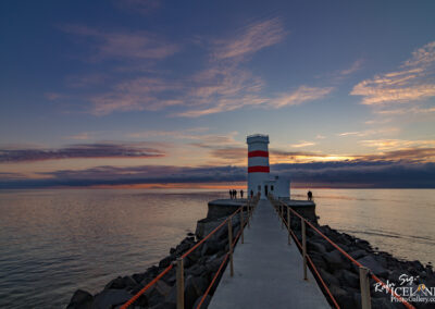 A lighthouse with alternating red and white stripes stands at the end of a concrete pier extending over calm waters. The sky is painted with shades of blue and orange as the sun sets, creating a beautiful sunset backdrop. Several figures can be seen on the pier, enjoying the view, while rocky formations line the water's edge.