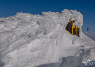 A snowy landscape featuring a wooden structure partially covered in thick layers of snow. The structure has yellow accents and a window, while a shovel leans against it. In the background, the sky is clear and blue, showcasing the contrast between the whiteness of the snow and the bright sky. The snow appears textured with waves and ridges, emphasizing the cold and wintery environment.