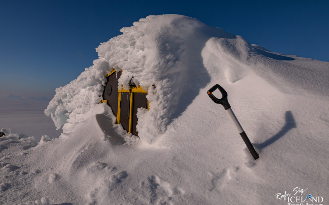 A small building partially buried in snow, with a yellow and brown exterior. The roof is covered in thick layers of snow and ice, while a black and white shovel stands upright nearby, partially buried as well. The background features a clear blue sky and the vast, white snowy landscape extending into the distance.