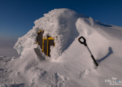 A small building partially buried in snow, with a yellow and brown exterior. The roof is covered in thick layers of snow and ice, while a black and white shovel stands upright nearby, partially buried as well. The background features a clear blue sky and the vast, white snowy landscape extending into the distance.