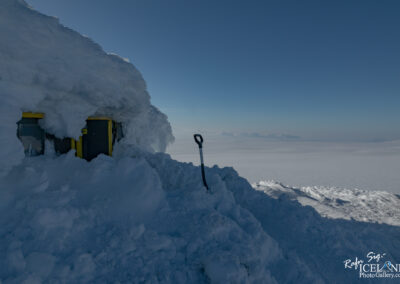 A snow-covered structure with yellow windows and a dark entrance is partially buried under a thick layer of snow. A snow shovel stands upright in the foreground, surrounded by deep snow. In the background, a vast, flat expanse of snow and ice stretches to the horizon under a clear blue sky.