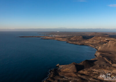 A panoramic view of a rugged coastline featuring dark hills and cliffs, with a clear blue sky above. The sea is calm, reflecting the sky, and in the distance, small buildings can be seen near the shoreline, suggesting a settlement. The landscape is predominantly earthy tones, with some white patches likely indicating snow on the hills.