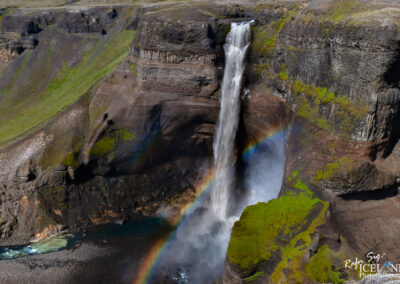 A stunning waterfall cascades down a rocky cliff with lush green vegetation on either side. At the base of the waterfall, a splash creates mist, and a vibrant rainbow arcs through the spray, adding color to the rugged landscape. The surrounding terrain features a mix of dark rocks and greenery, with a clear blue river flowing near the bottom. The scene captures the beauty and power of nature in a serene setting.