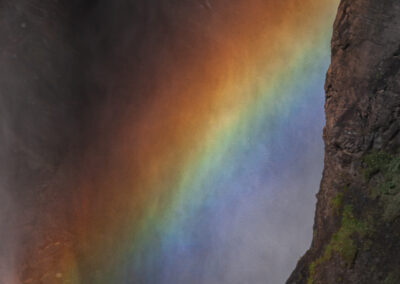 A vibrant rainbow arcs across a waterfall, set against a backdrop of dark rock surfaces. The colors transition smoothly from deep orange to bright red, continuing into green and blue hues. Mist from the waterfall partially obscures the scene, adding a soft glow to the colors.