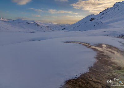 A tranquil winter landscape featuring a vast expanse of snow-covered terrain, rolling hills, and distant mountains under a partly cloudy sky. The scene includes a small area of exposed ground with a reflective surface, likely water, creating a subtle contrast against the white snow. The soft light adds a serene ambiance to the picturesque view, highlighting the natural beauty of the icy landscape.
