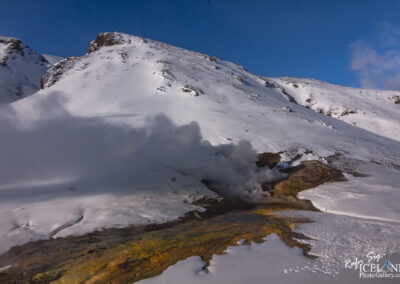 A snowy landscape featuring a mountainous terrain with a prominent peak. In the foreground, there is a geothermal area emitting steam, surrounded by a patch of colorful mineral-rich ground. The scene is set against a clear blue sky, enhancing the contrast between the white snow and the vibrant earth tones below.