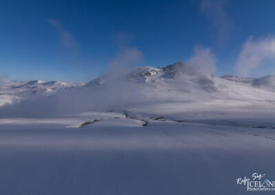 A vast, snow-covered landscape under a clear blue sky, featuring gentle rolling hills and distant mountains. Wisps of fog or steam rise from the terrain, adding a mystical quality to the scene. The ground shows smooth, untouched snow interspersed with subtle crevices, highlighting the serene isolation of the environment.
