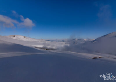 A vast, snow-covered landscape stretches across the scene, featuring rolling hills and gentle slopes under a clear blue sky. Wisps of clouds float above the terrain, and faint wisps of steam emerge from a distant area, hinting at geothermal activity. The snow glistens under the sunlight, creating a serene and pristine atmosphere.