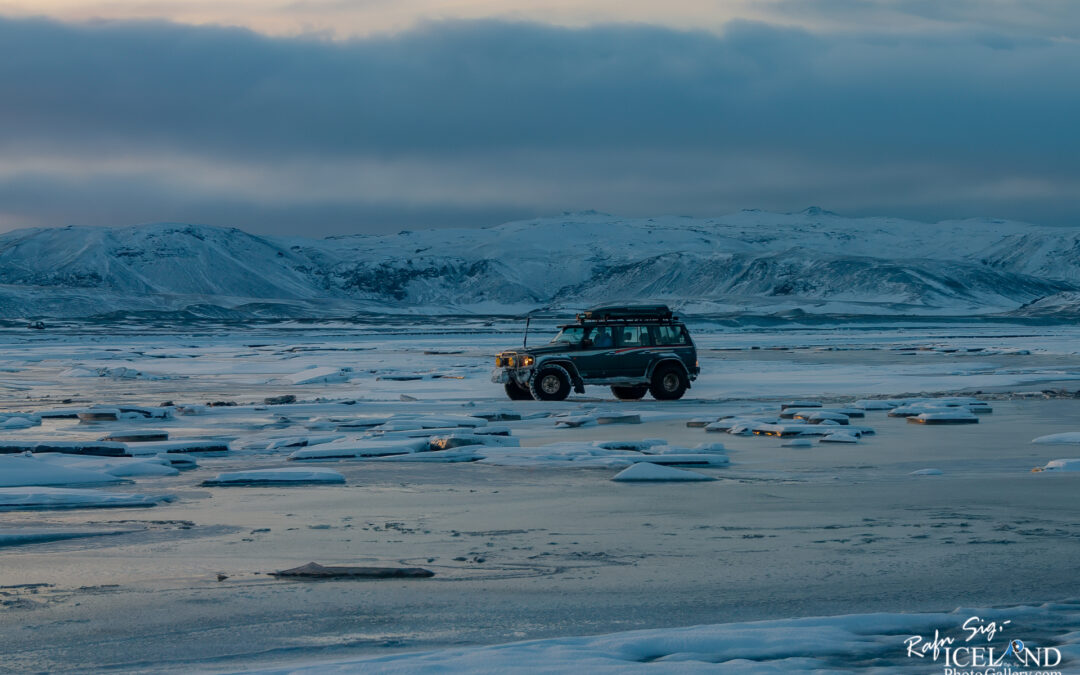 A four-wheel-drive vehicle is parked on a frozen landscape scattered with ice floes. The background features snow-covered mountains under a cloudy sky, with soft light from the setting sun illuminating the scene. The icy terrain reflects the surrounding colors, creating a serene and remote atmosphere.