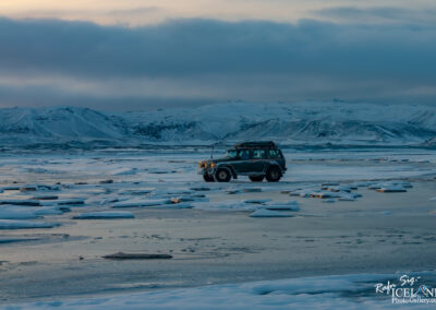 A four-wheel-drive vehicle is parked on a frozen landscape scattered with ice floes. The background features snow-covered mountains under a cloudy sky, with soft light from the setting sun illuminating the scene. The icy terrain reflects the surrounding colors, creating a serene and remote atmosphere.