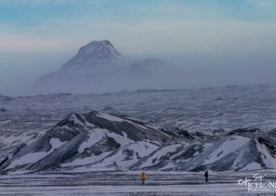 A vast, icy landscape featuring rolling hills and mountains covered in snow. In the foreground, two small men are visible, one wearing a bright yellow jacket and the other dressed in darker clothing. In the background, a large, snow-capped mountain looms through a misty atmosphere, giving a sense of depth and scale to the scene. The sky above is pale blue, transitioning from light to darker shades, enhancing the serene yet dramatic ambiance of the cold environment