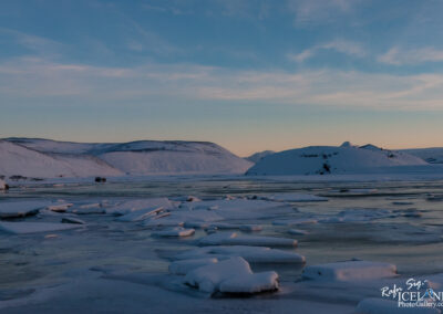 A serene winter landscape featuring a tranquil body of water surrounded by icy shores. Ice floes are scattered across the water's surface, while snow-covered hills rise in the background under a softly lit sky at dusk. The colors range from light blue to soft pastel hues, creating a peaceful and picturesque winter scene.