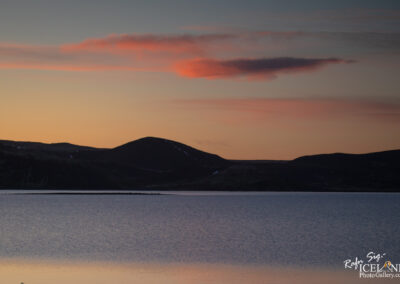 A serene landscape at twilight featuring a calm lake that reflects the soft colors of the sky. In the background, dark silhouettes of hills and mountains rise against a gradient of blues and oranges, while wispy clouds are tinted pink and orange. The scene conveys a peaceful and tranquil atmosphere.