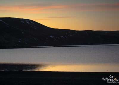 A tranquil lake reflecting the surrounding mountains at sunset, with soft hues of orange and pink in the sky. The mountains are partially covered in snow, and the water's surface displays gentle ripples, adding to the serene atmosphere of the landscape.