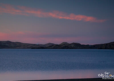 A tranquil lake surrounded by rolling hills and mountains, reflecting a soft, pastel-colored sky at dusk. The water appears calm, with gentle ripples, while wispy clouds with hints of pink and orange contrast against the fading light in the background.