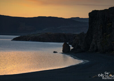 A serene landscape featuring a calm body of water reflecting hues of orange and gold from a sunset. The shore is lined with dark, rocky formations, and a gentle curve of black sand is visible. In the background, rolling hills rise against the dusk sky, creating a tranquil and picturesque scene.