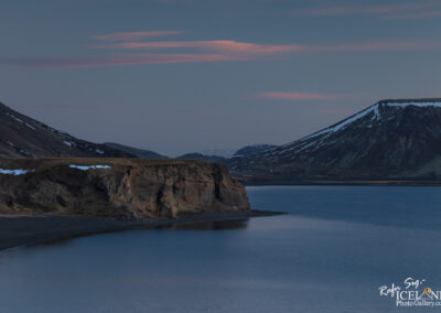 A serene landscape featuring a calm body of water bordered by rocky cliffs and snow-capped mountains in the distance. The scene is bathed in soft twilight, with gentle pink clouds in the sky above. The terrain is rugged and natural, with a hint of vegetation and exposed rock.