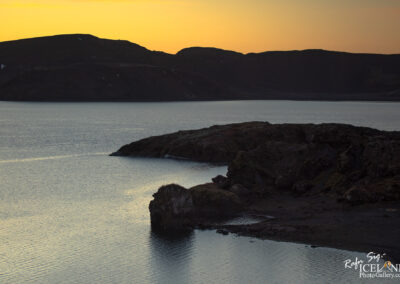 A serene landscape at twilight featuring a calm lake reflecting soft hues of orange and yellow from the sunset. Dark, rugged hills frame the background, while a rocky outcrop juts into the lake from the foreground, adding a natural element to the tranquil scene.