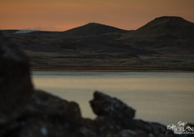A tranquil landscape featuring a calm body of water reflecting the colors of the sunset. In the background, gently rolling hills and mountains are visible, with some areas appearing dark and others covered in green patches. A plume of steam rises from a distant area, hinting at geothermal activity. In the foreground, rocky formations frame the scene, adding depth to the composition.
