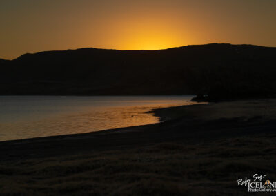 A serene landscape at dusk, showcasing a calm lake reflecting warm hues of orange and yellow from the setting sun. The silhouette of a dark mountain range borders the horizon, contrasting with the vibrant sky, while the shoreline is defined by gentle waves and patches of grass. The scene evokes a sense of tranquility and natural beauty.