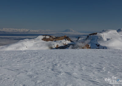 A vast, snowy landscape stretches across the foreground, with gentle rolling hills and mountains in the background. The scene is illuminated by clear blue skies, showcasing a peaceful winter day. Some rocky outcrops peek through the snow near the center, while steam rises from an area on the left, hinting at geothermal activity. In the distance, snow-capped mountains are visible, creating a stunning panoramic view of the rugged terrain.