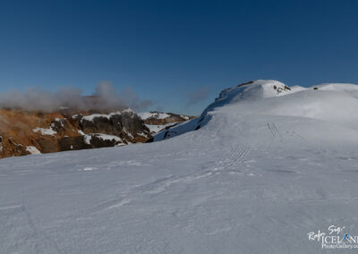 A snowy landscape blanketed by fresh snow, with a clear blue sky overhead. In the background, rugged rocky formations with patches of snow contrast against the earth tones of the rocks. Wispy clouds drift above the mountains, creating a serene winter scene. Snowmobile tracks are visible on the smooth white surface, leading toward the distant hills.