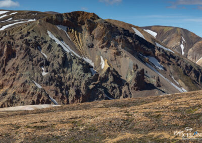A panoramic view of rugged mountain terrain showcasing a mix of earthy tones including browns, greens, and yellows. The landscape features steep, rocky slopes with patches of snow and exposed rock faces, set against a clear blue sky. The foreground consists of a mix of gravel and sparse vegetation. The overall scene conveys a remote and natural wilderness area, likely in a volcanic region.