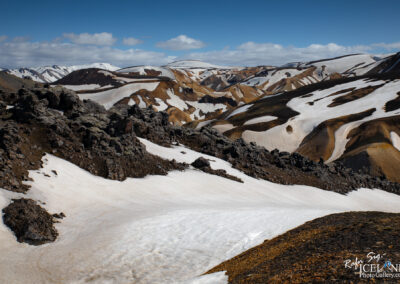 A panoramic view of a mountainous landscape featuring a mix of snow-capped peaks, rocky outcrops, and vibrant patches of soil in shades of brown and orange. The scene is set under a clear blue sky, with fluffy white clouds scattered throughout. The contrasting textures of the snow and rocky terrain create a striking visual effect, showcasing the natural beauty of the environment.