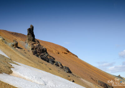 A rocky landscape with a prominent, tall rock formation on a slope. The terrain is primarily composed of golden-brown earth and scattered stones, with a small patch of snow in the foreground. The sky above transitions from light blue to deeper hues, indicating a clear day. The rock formation appears rugged and jagged, contrasting with the smoother gradient of the slope.
